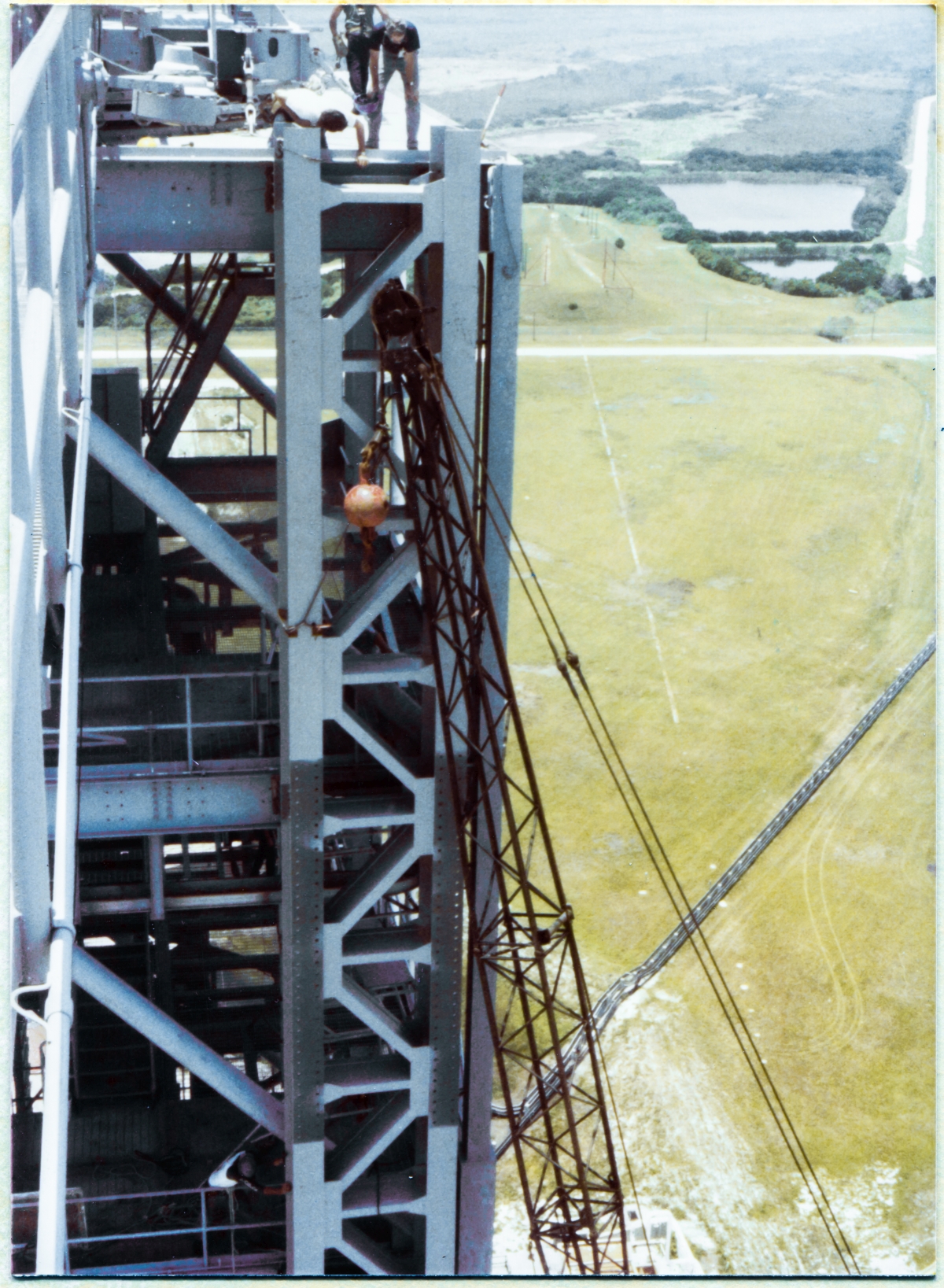 Image 084. Wade Ivey (dark blue shirt, holding blue hardhat in his right hand), owner of Ivey's Steel Erectors, stands on the checkerplate which defines the top of the Fixed Service Structure at elevation 300'-0”, in this image taken from the far end of the Hammerhead Crane Boom at Space Shuttle Launch Complex 39-B, Kennedy Space Center, Florida, and leans out past the edge of the checkerplate in order to see precisely where his team of Union Ironworkers from Local 808 have positioned the GOX Arm Hinges Support Strongback. The Strongback is not quite at its final location, where it will be welded and bolted to the Perimeter Framing of the FSS, and great care must be taken using a mobile crane with just under 250 feet of boom, maneuvering horizontally and vertically to sub-eighth-inch tolerance, in conjunction with rigging attached directly to the FSS itself, to get the Strongback EXACTLY where it belongs, where it will be fastened immovably in place, thus irrevocably locating where the GOX Arm itself will be positioned for its servicing work at the Gaseous Oxygen Vent on top of the Space Shuttle's External Tank. This is exceedingly close-tolerance work, and it takes the best in the business to execute it, and Wade Ivey along with his whole workforce of Union Ironworkers were the best in the business. Photo by James MacLaren.